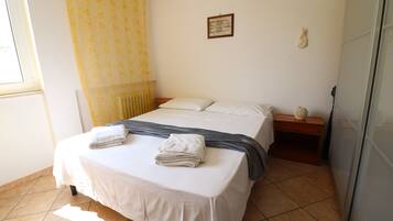 A neatly made bed with white linens and a gray blanket, a small bedside table with a lamp, and a window with yellow curtains.