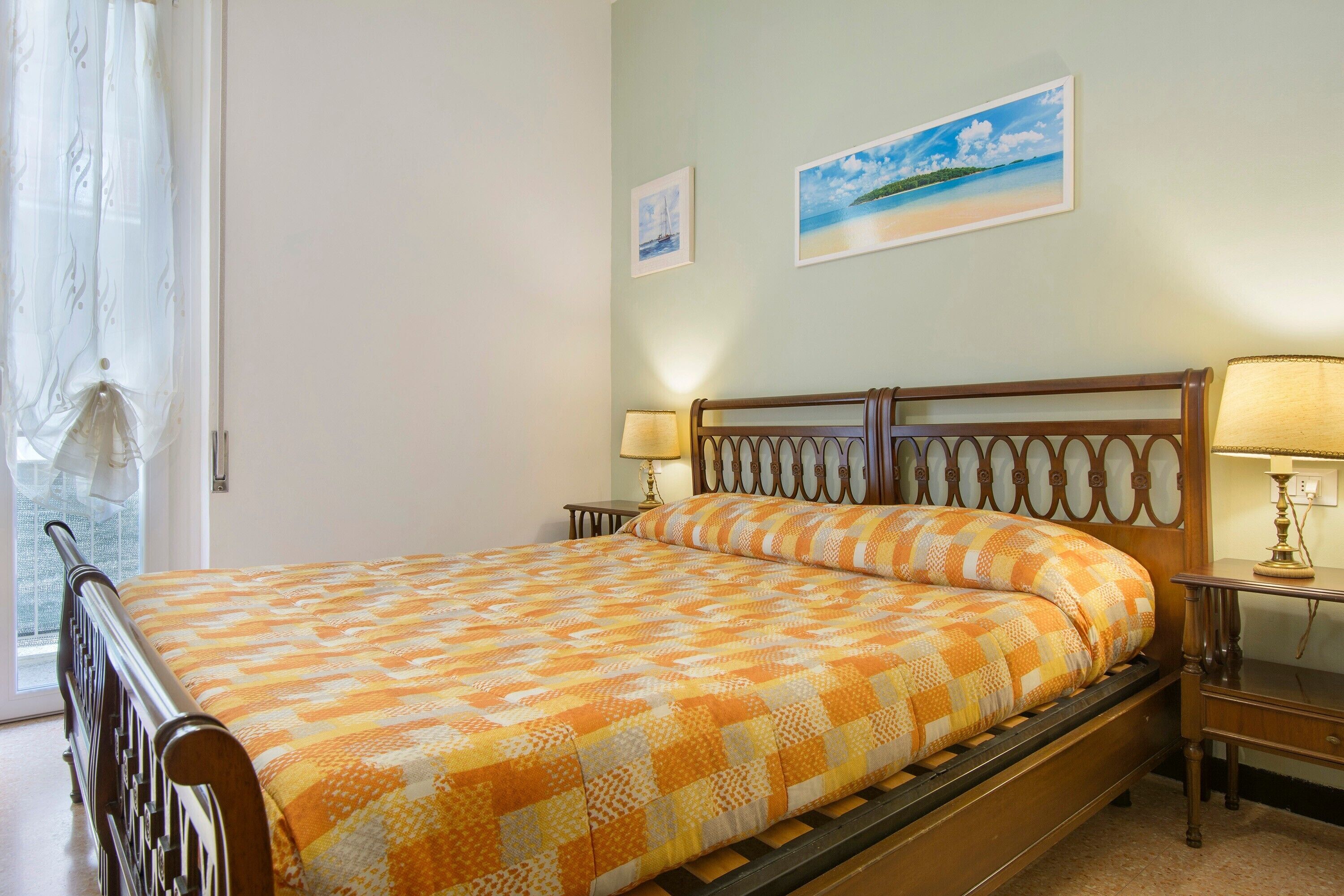 Una cama con una colcha estampada, un cabecero de madera, dos mesitas de noche con lámparas y una ventana con cortinas.
