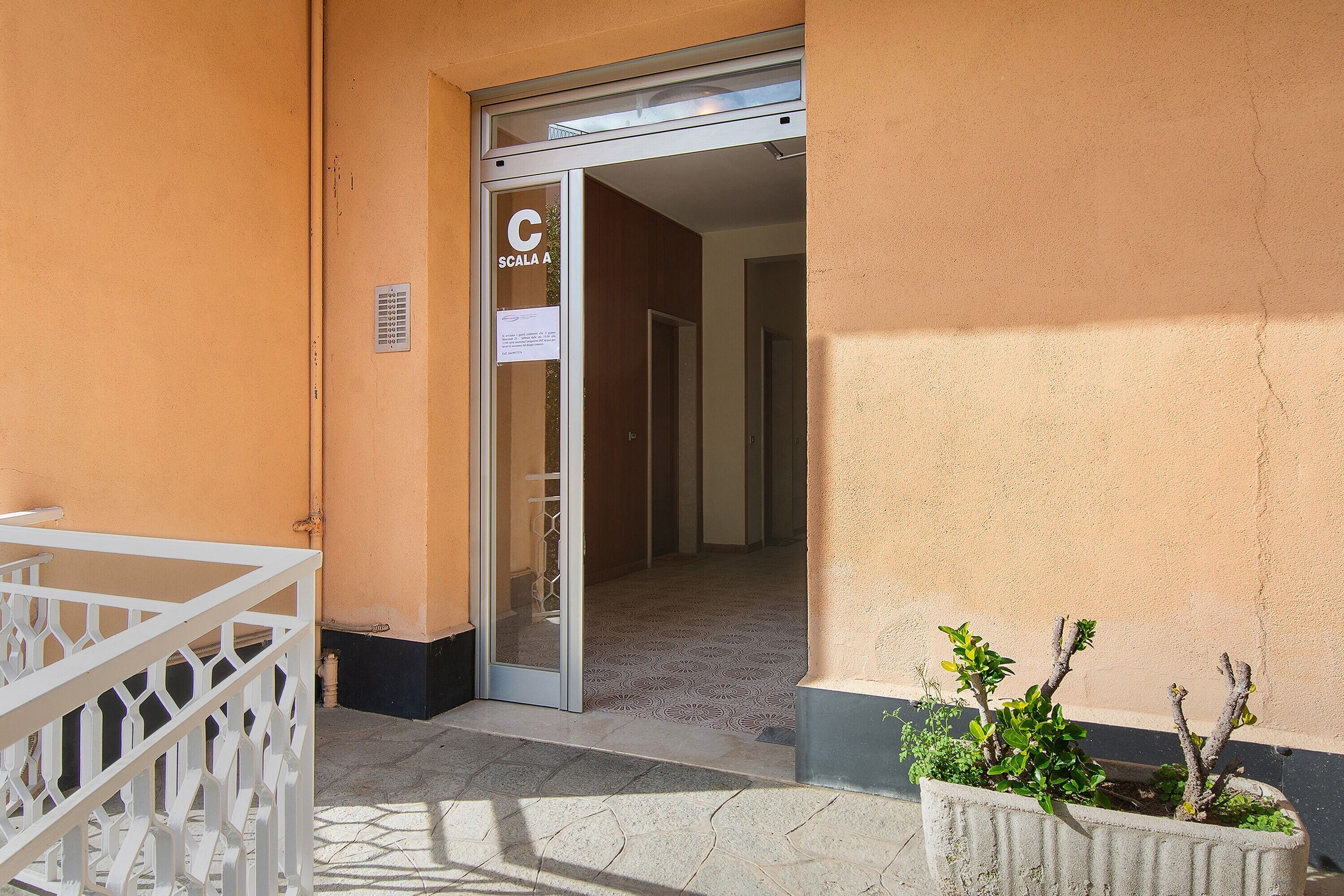 Ingresso di un edificio con una porta in vetro che reca la scritta 'C SCALA A', una ringhiera in metallo bianco e un vaso con piante verdi.