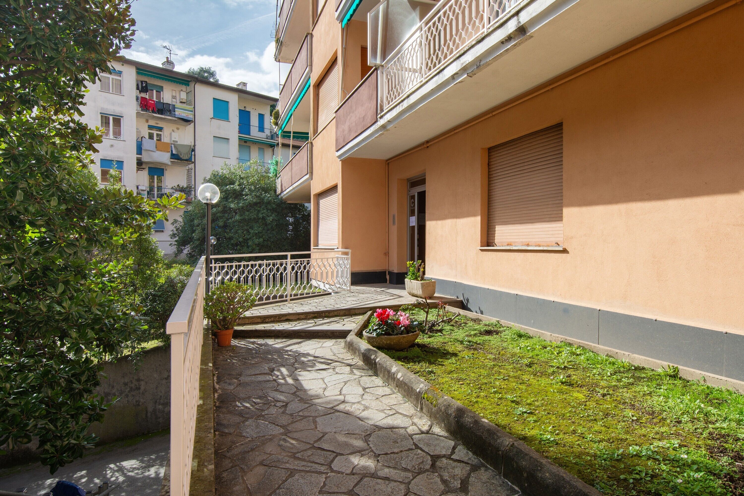 A residential building with a stone pathway, a small garden with flowers, and a balcony with a railing.