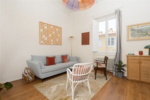 A living room with a light blue sofa, a white wicker chair, a floor lamp, and a large window with curtains. - San Frediano Designer Duplex (Firenze)