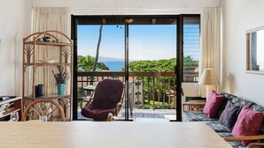 Una habitación con una silla de madera, un sofá con cojines, una mesa y vista al océano a través de un ventanal corredero.