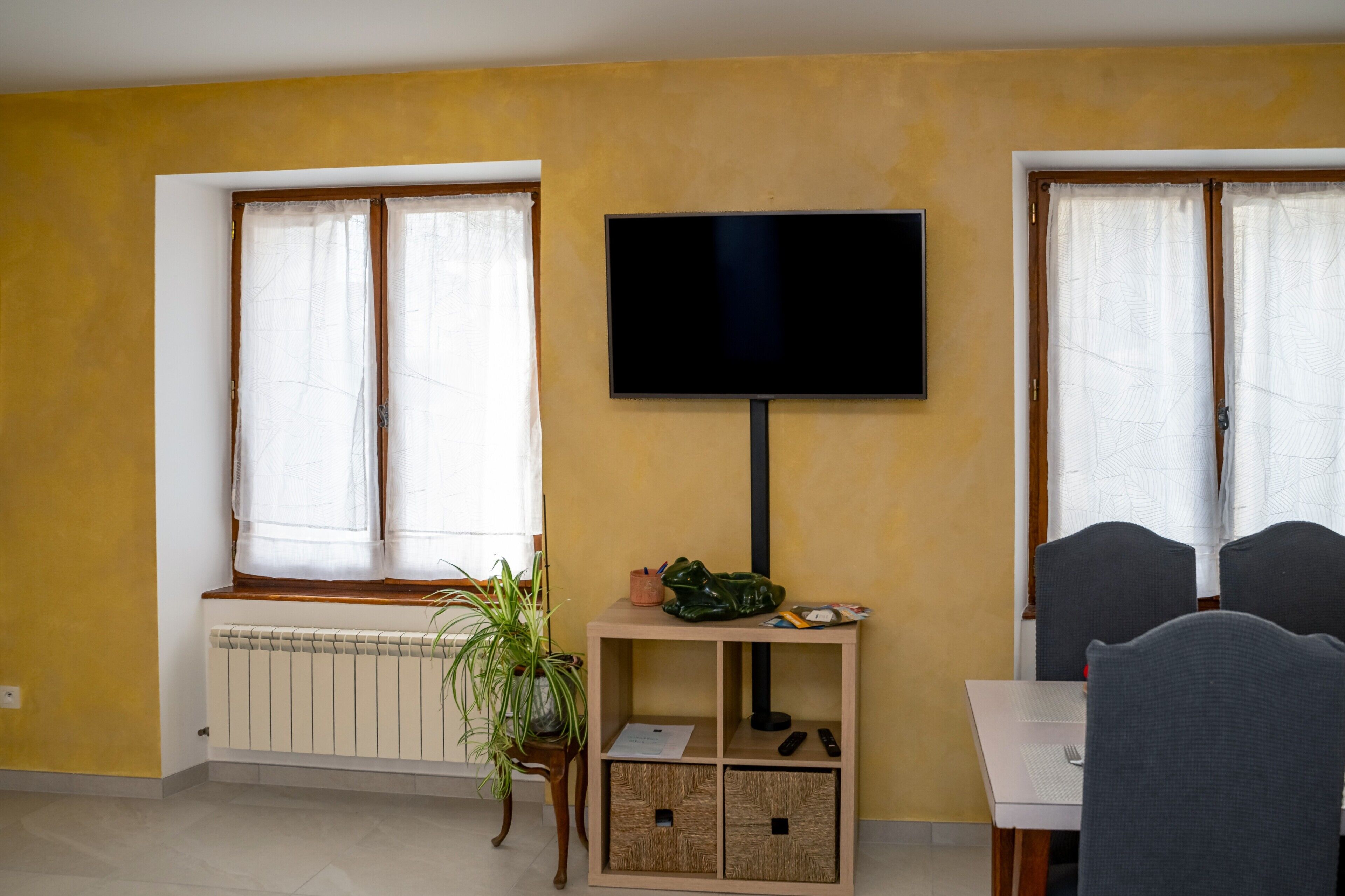 A room with a TV mounted on the wall, a radiator, a window with curtains, a small table with a plant, and a dining area with chairs.