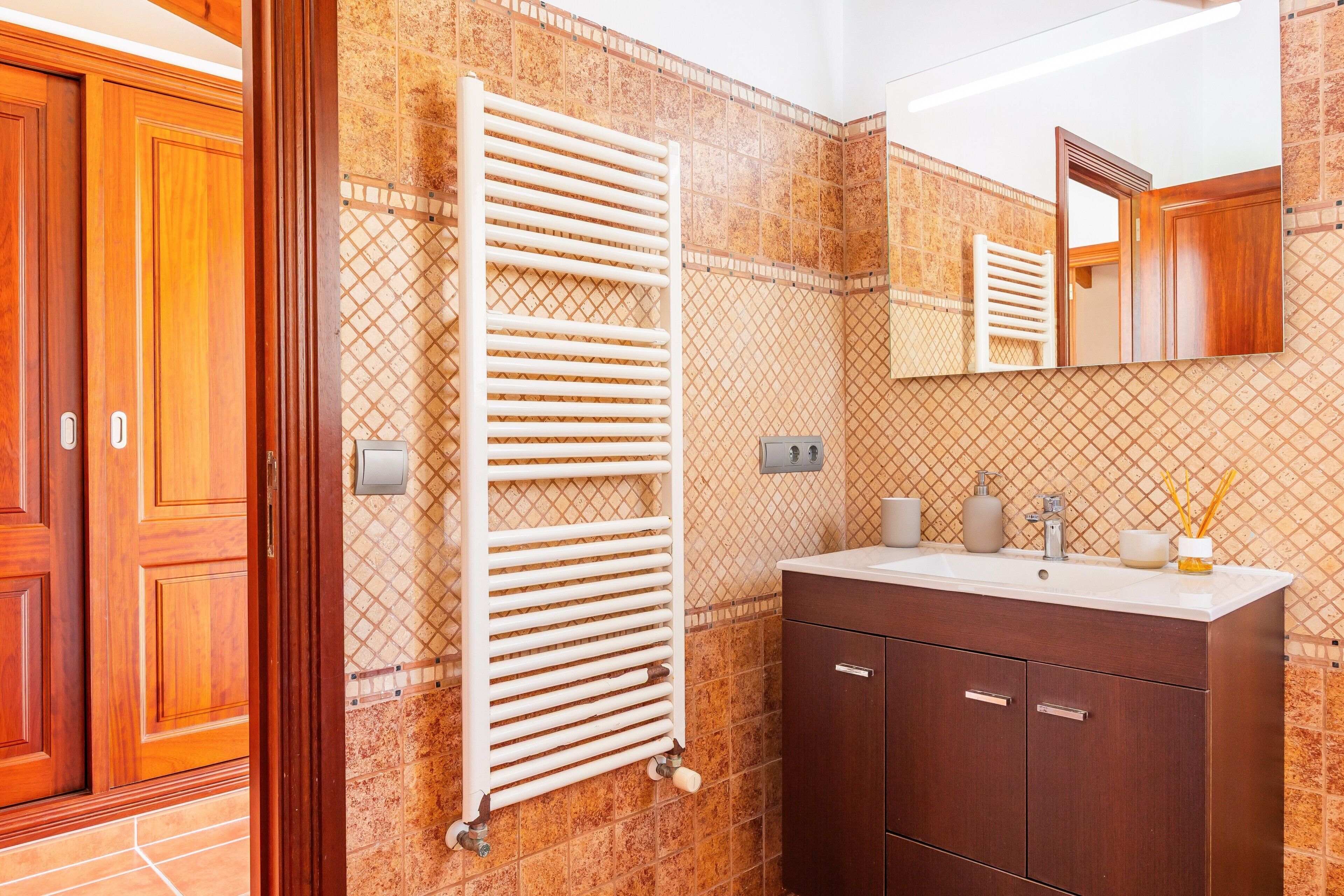 Un baño con puerta de madera, toallero blanco, lavabo con artículos de aseo y espejo.