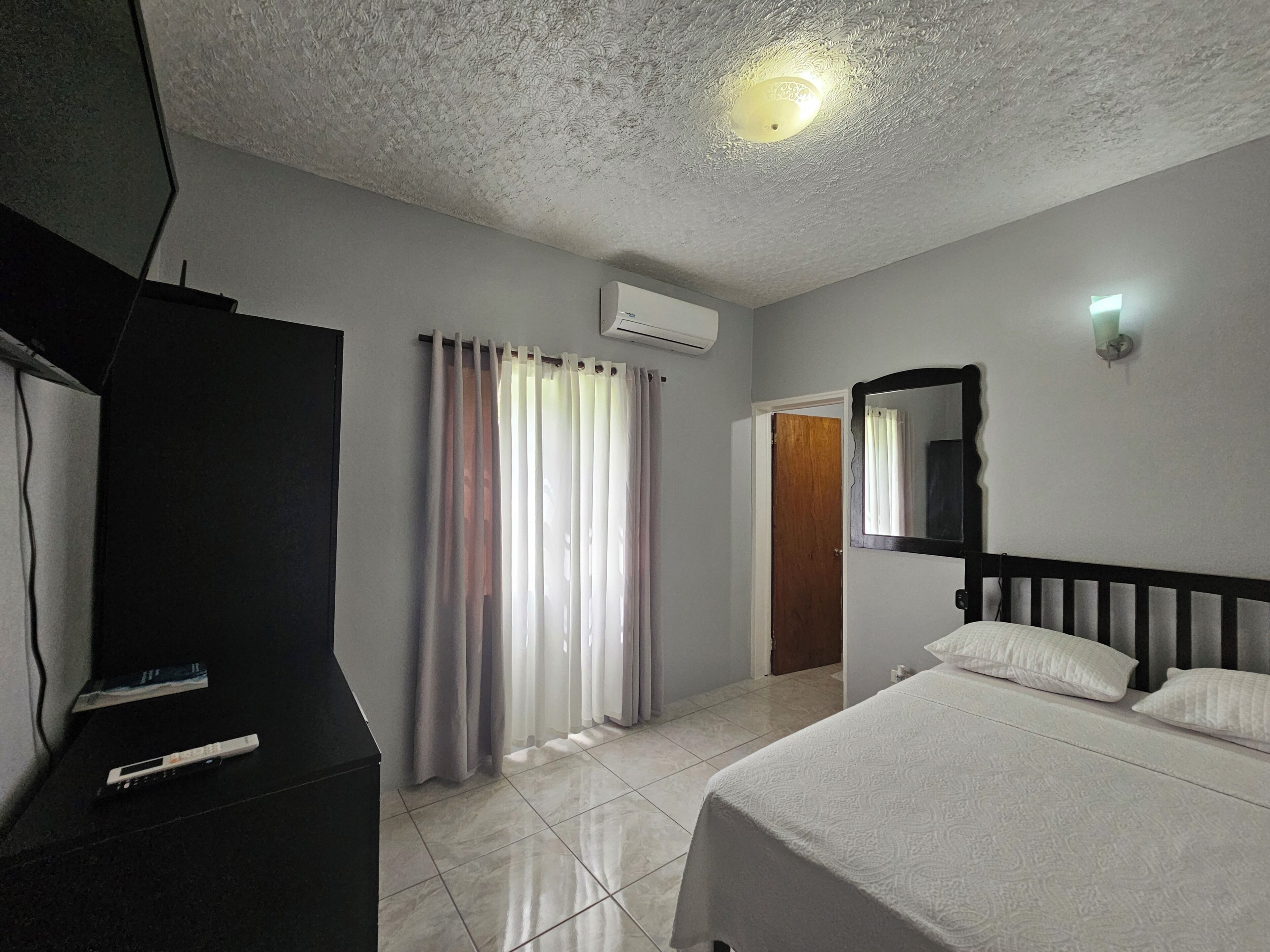 A hotel room with a bed, a television, curtains, a mirror, and a ceiling light.