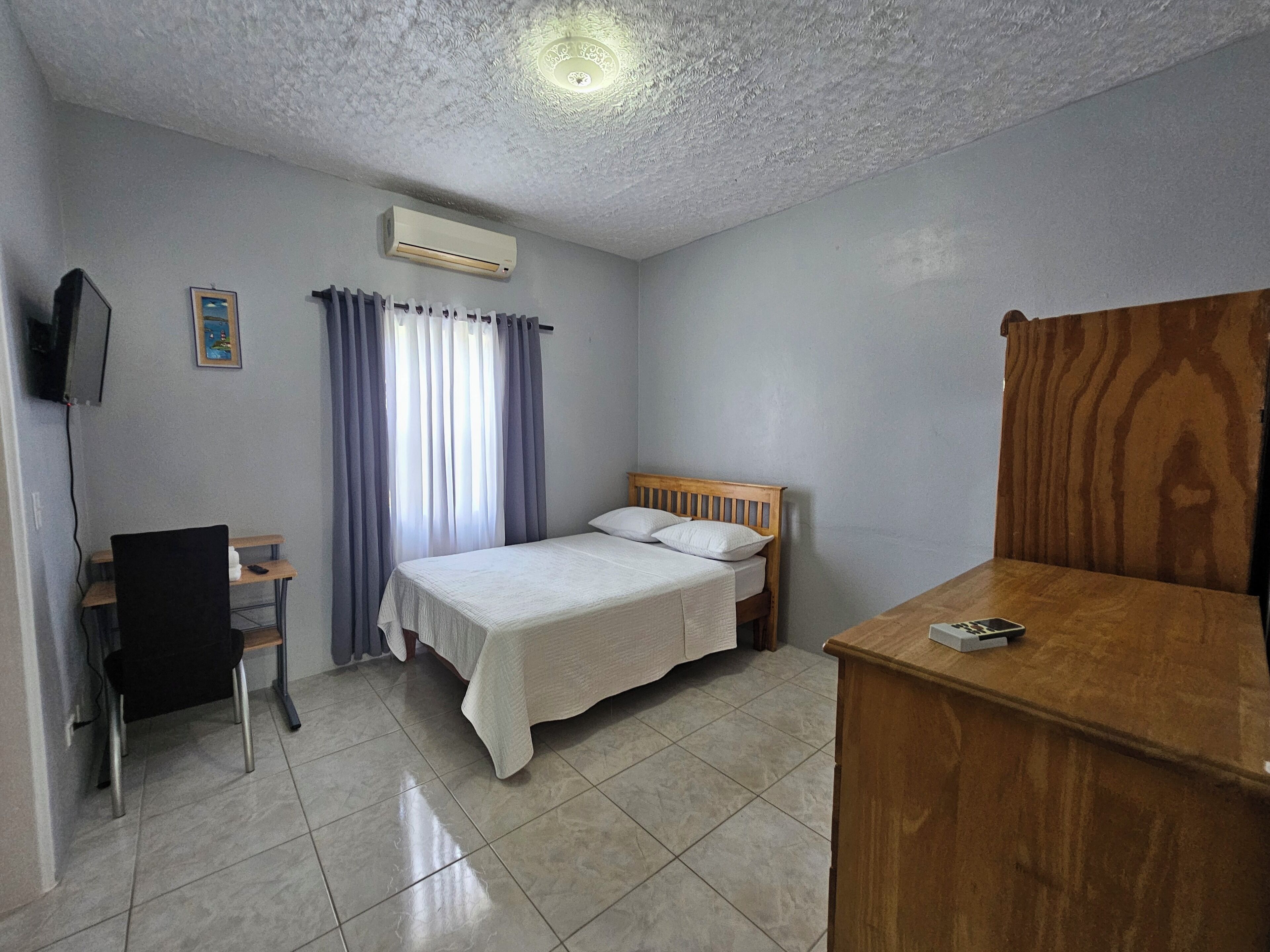A bedroom with a bed, a desk, a chair, an air conditioner, and a television mounted on the wall.