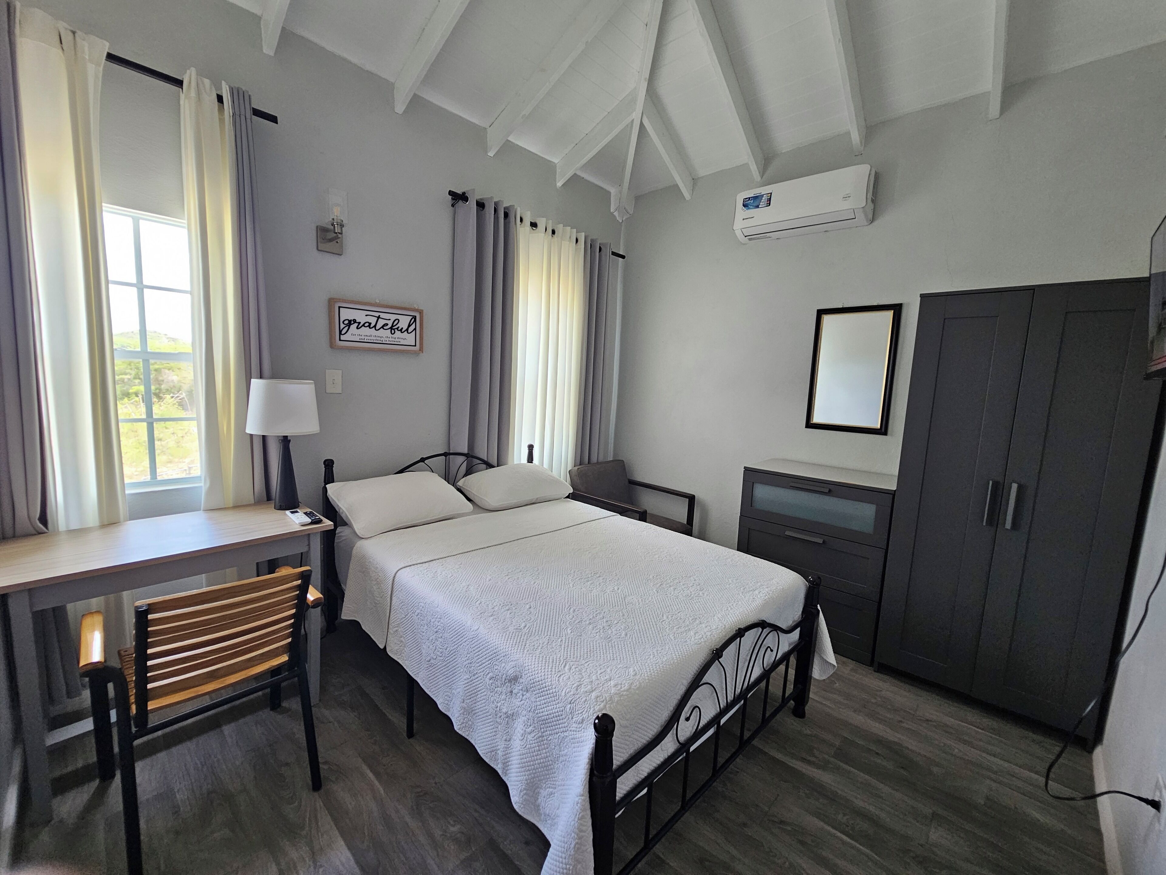 Un dormitorio con cama, escritorio, silla, armario y ventana con cortinas.
