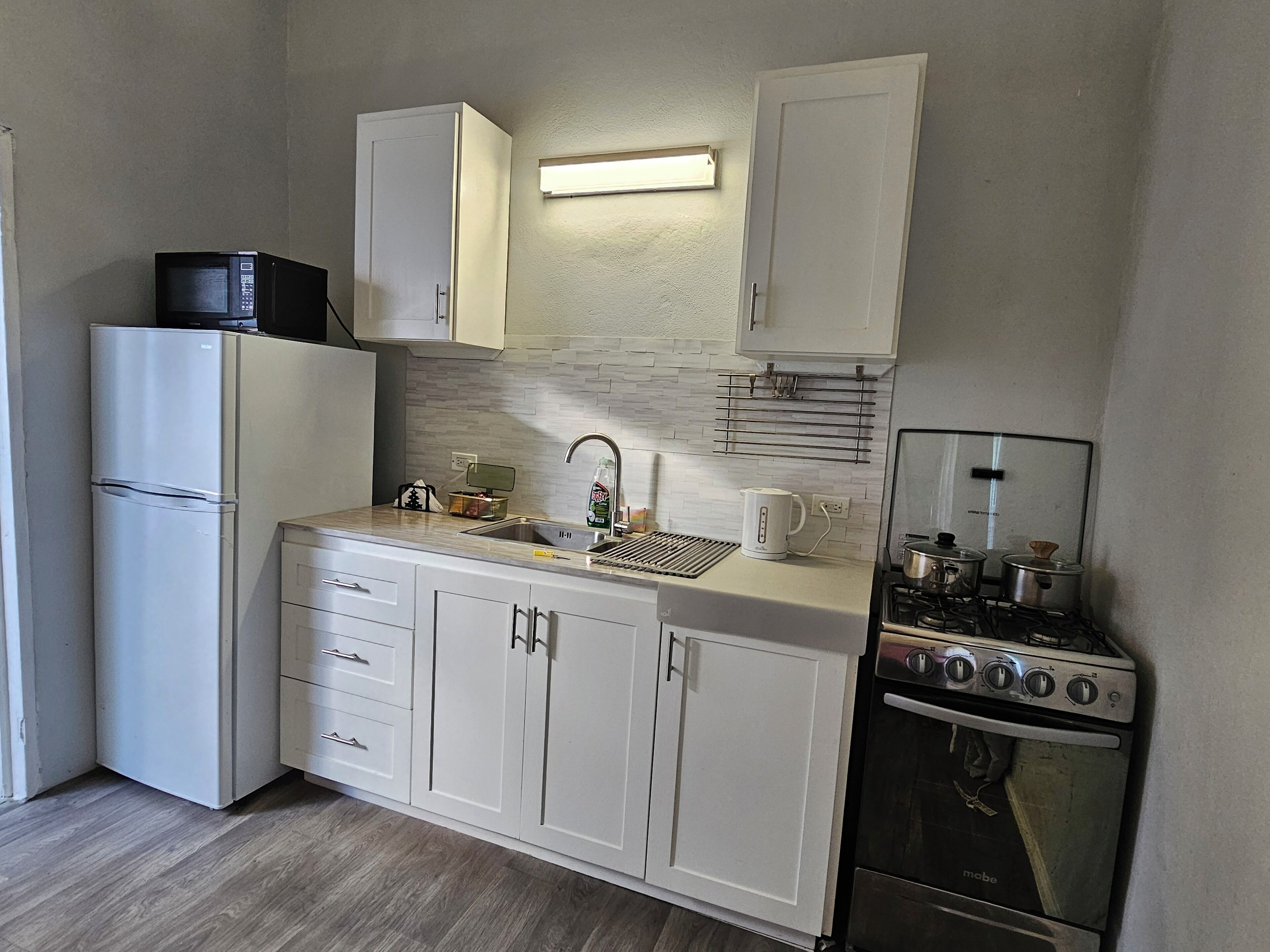 A compact kitchen with white cabinets, a stainless steel sink, a microwave, a kettle, and a stove.
