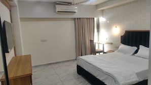 A hotel room with a bed, a desk, an air conditioner, a chair, and a window with curtains. - Januson Hotel (Gandhinagar)