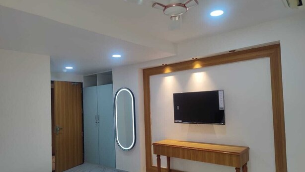 A bedroom with a bed, a wooden bedside table, a TV mounted on the wall, a wardrobe, and a mirror. - Januson Hotel (Gandhinagar)