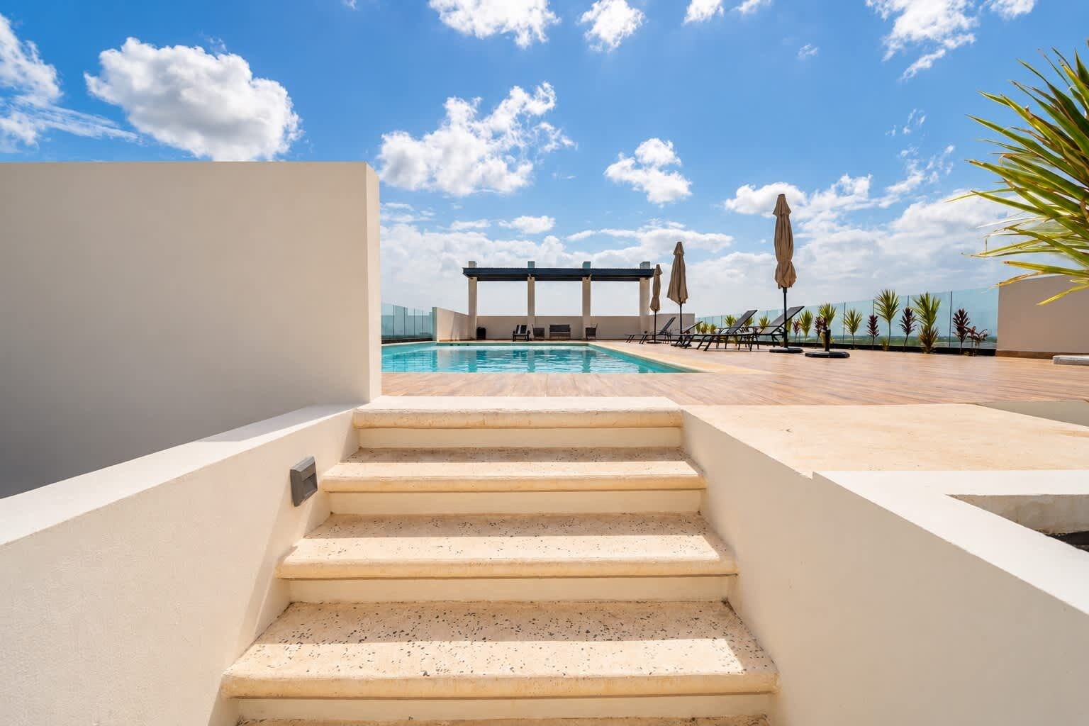 A pool area with steps leading to a covered seating area, surrounded by lounge chairs and umbrellas.