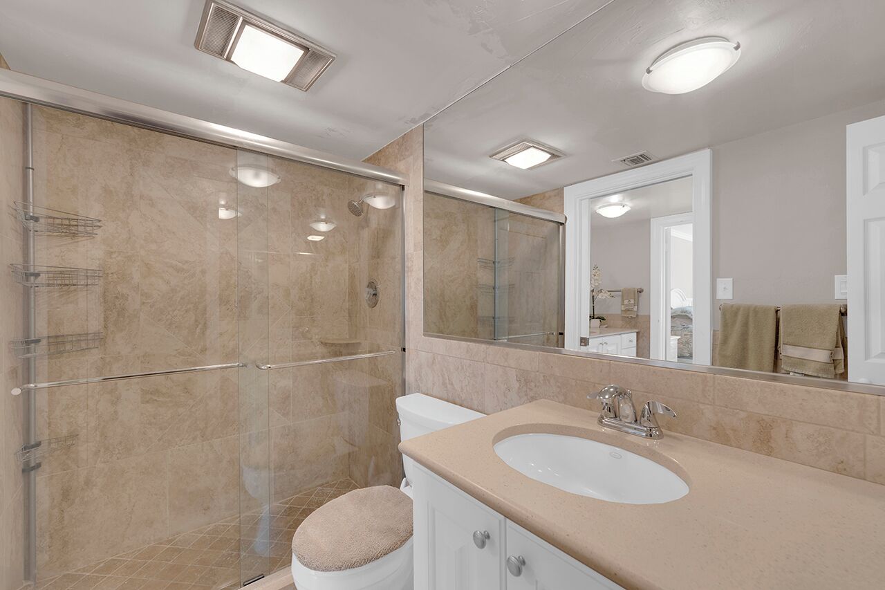 A modern bathroom with a glass shower enclosure, a white toilet, a beige sink, and a large mirror.