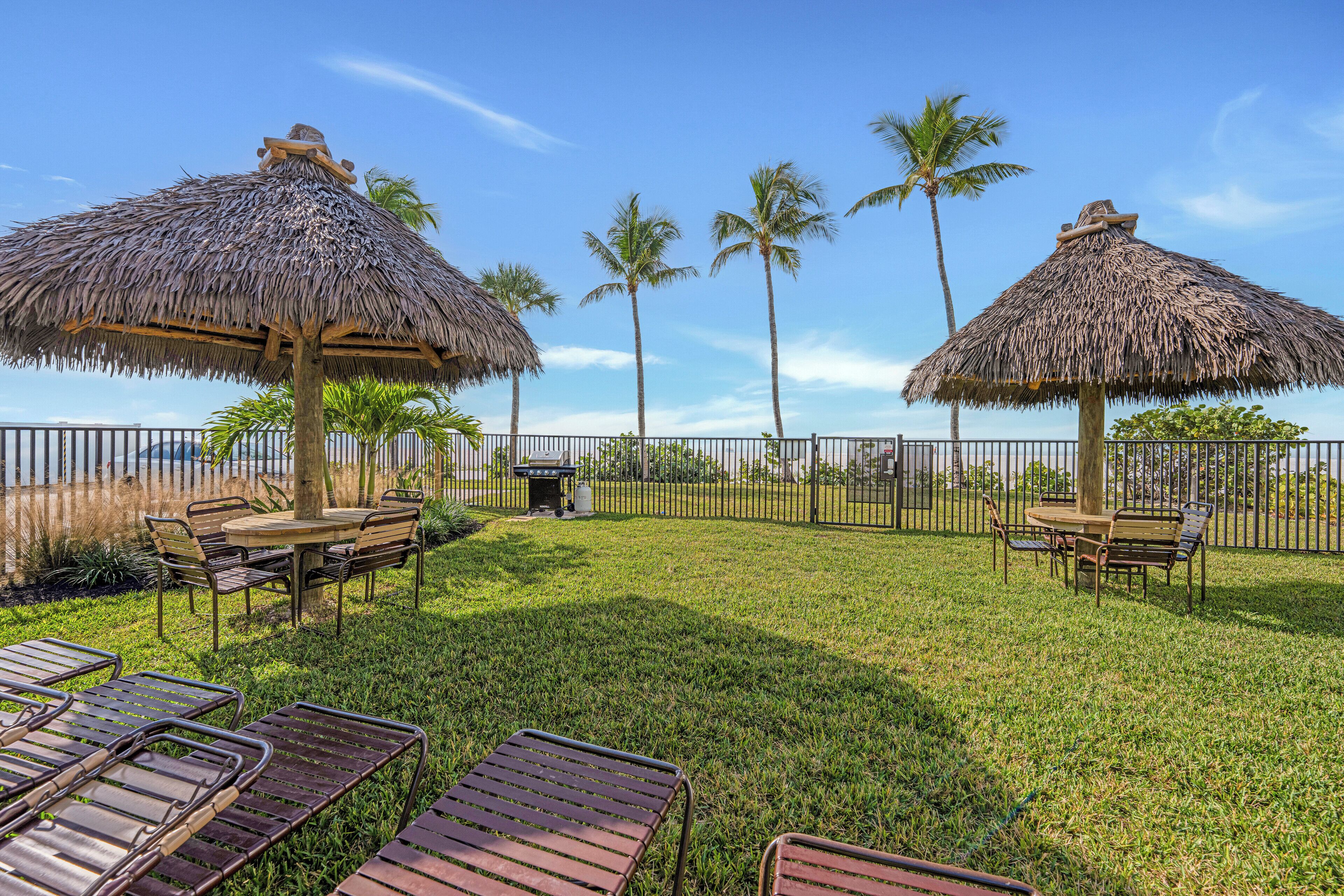 A tropical outdoor seating area with thatched umbrellas, metal chairs, and a table, surrounded by a fence and palm trees.