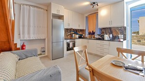 Cocina moderna con armarios blancos, un refrigerador de acero inoxidable y un horno empotrado. Hay un comedor con una mesa y sillas de madera, y vistas al exterior a través de un amplio ventanal.