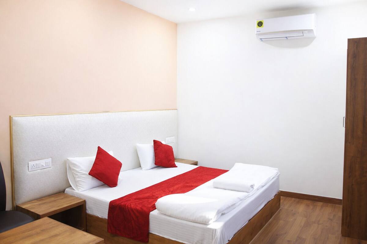 A hotel room with a bed, a nightstand, a wooden floor, a white wall, and an air conditioning unit.