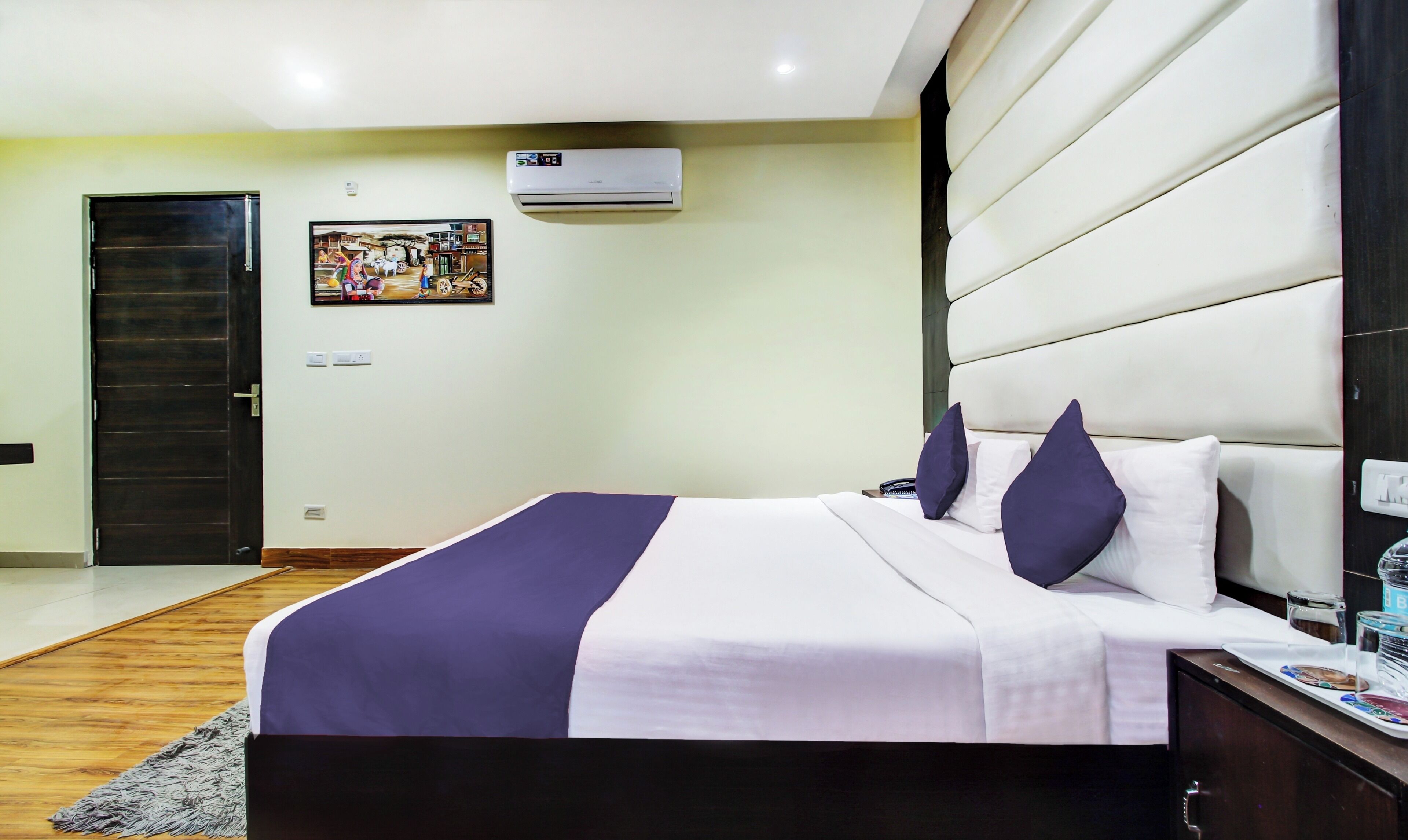A hotel room with a bed, a bedside table, a nightstand, a wall-mounted air conditioner, and a door.