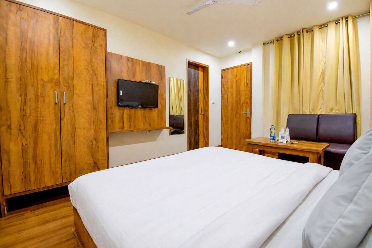 Quarto de hotel com uma cama grande, guarda-roupa de madeira, televisão, uma mesinha com cadeira e cortinas.