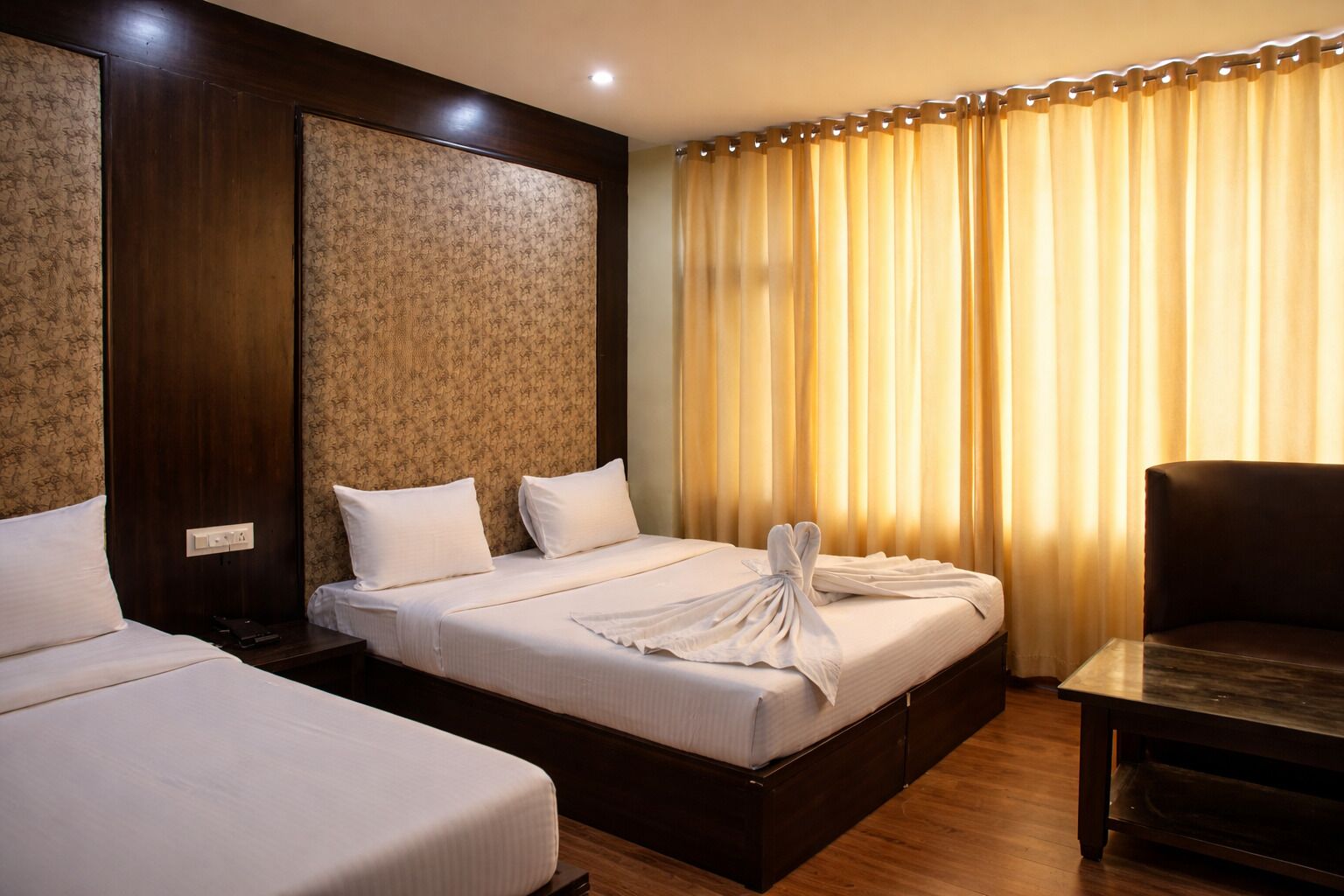 A hotel room with two beds, a wooden headboard, a wooden nightstand, a small table, and a window with curtains.