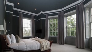 A large, dark-themed bedroom with a large bed, multiple windows, and a flat-screen TV mounted on the wall. - Frenchay Park House (Bristol)