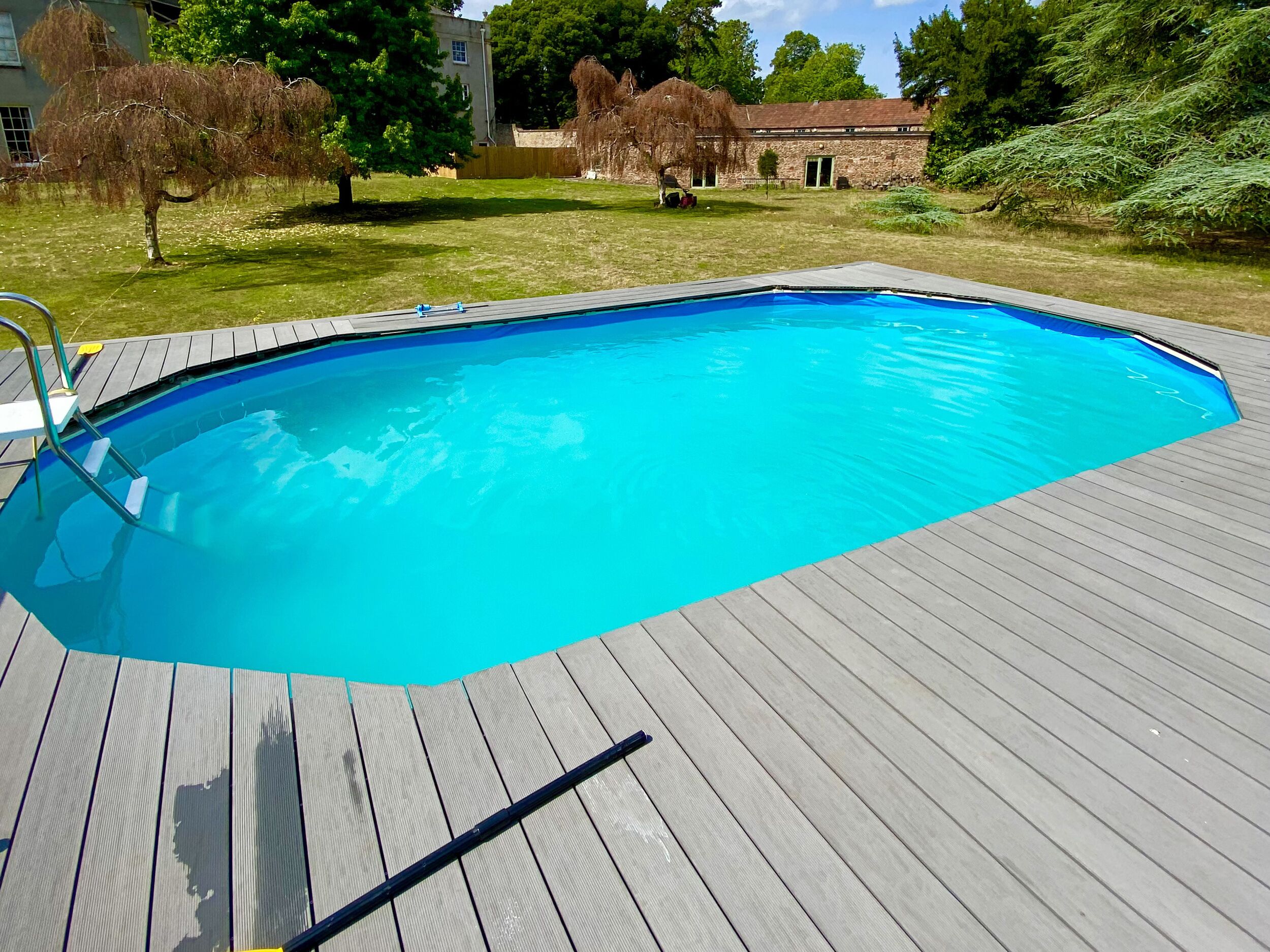 A rectangular swimming pool with a wooden deck, surrounded by a grassy yard and trees.
