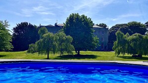 A swimming pool with a wooden deck in the foreground and a large building with trees in the background. - Frenchay Park House (Bristol)