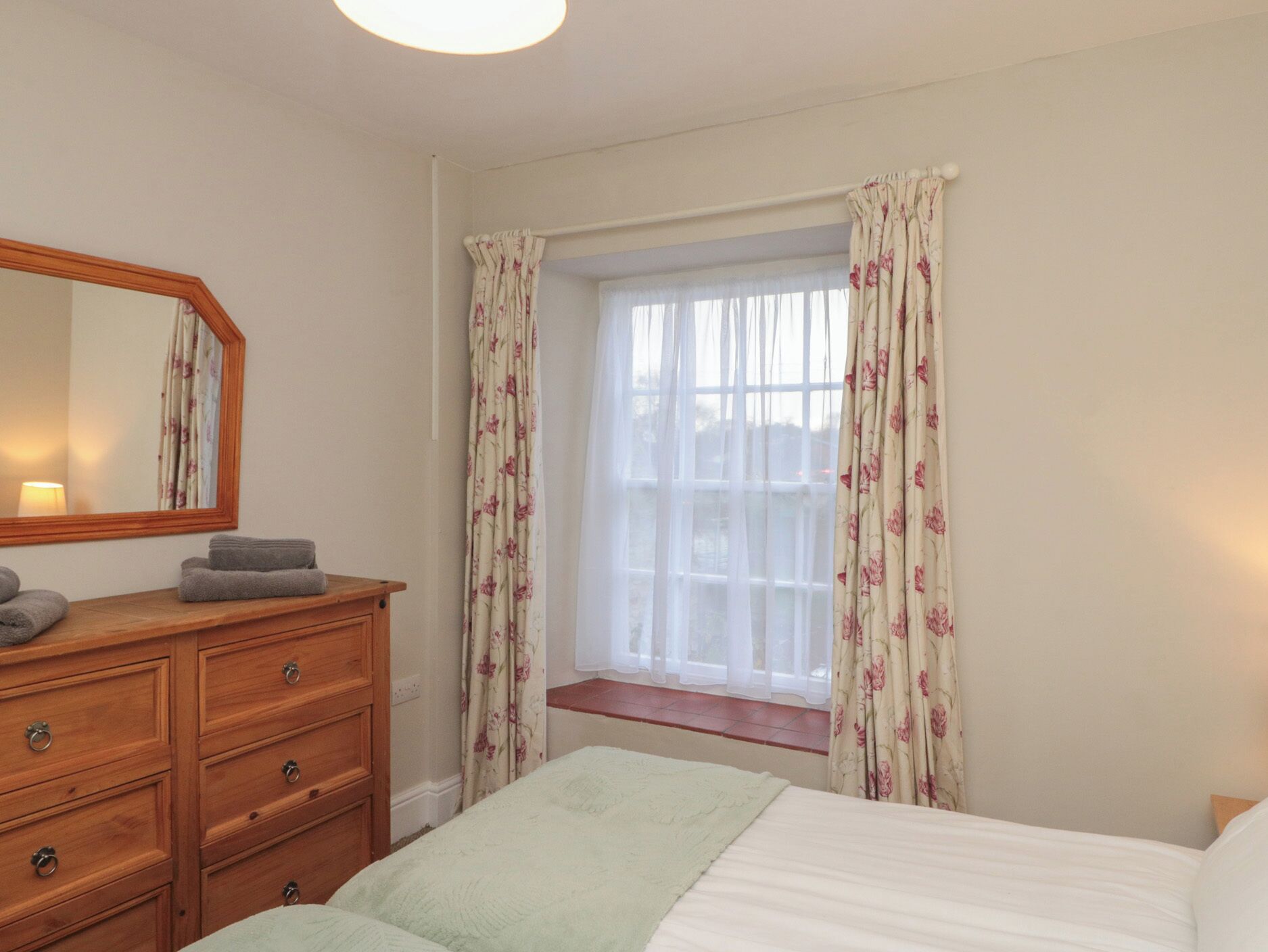 A bedroom with a wooden dresser, a bed with a green blanket, floral curtains, and a window with a view.