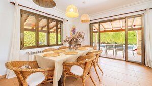 A dining area with a table set for six, wicker chairs, a vase with dried flowers, and large windows with a view of greenery. - Holiday Home 'Villa Cal Peso' with Mountain View, Wi-Fi and Air Conditioning (Illes Balears)
