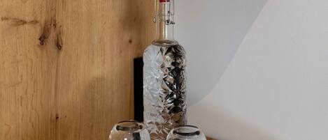 Una estantería de madera con una botella de vidrio, dos vasos y una rosa roja en una maceta verde.