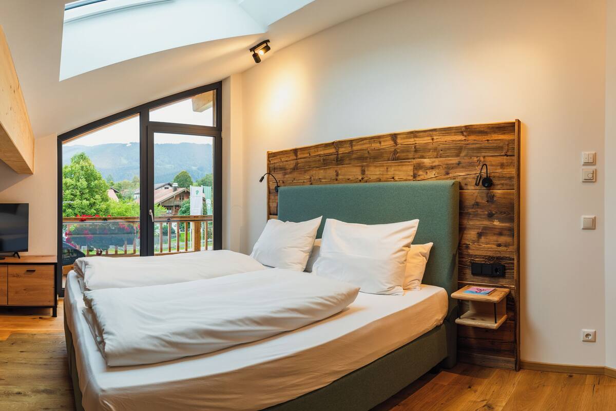 A bedroom with a large bed, a TV, and a view of a green landscape through a large window.