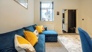 A modern living room with a blue sectional sofa, a white radiator, and a window with a view. - Stunning 3 Bedroom Home in Newcastle-under-lyme With Garden (Newcastle-Under-Lyme)