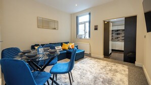 A modern living room with a dining area, featuring a sofa set, blue chairs, a glass-top table, and a large window providing natural light. - Cosy 4 Bed Townhouse Near Keele University and Royal Stoke Hospital (Newcastle-Under-Lyme)