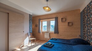 Ein Zimmer mit Holzwänden, einem großen Fenster, zwei Betten, einem Sessel und einer Pendelleuchte.