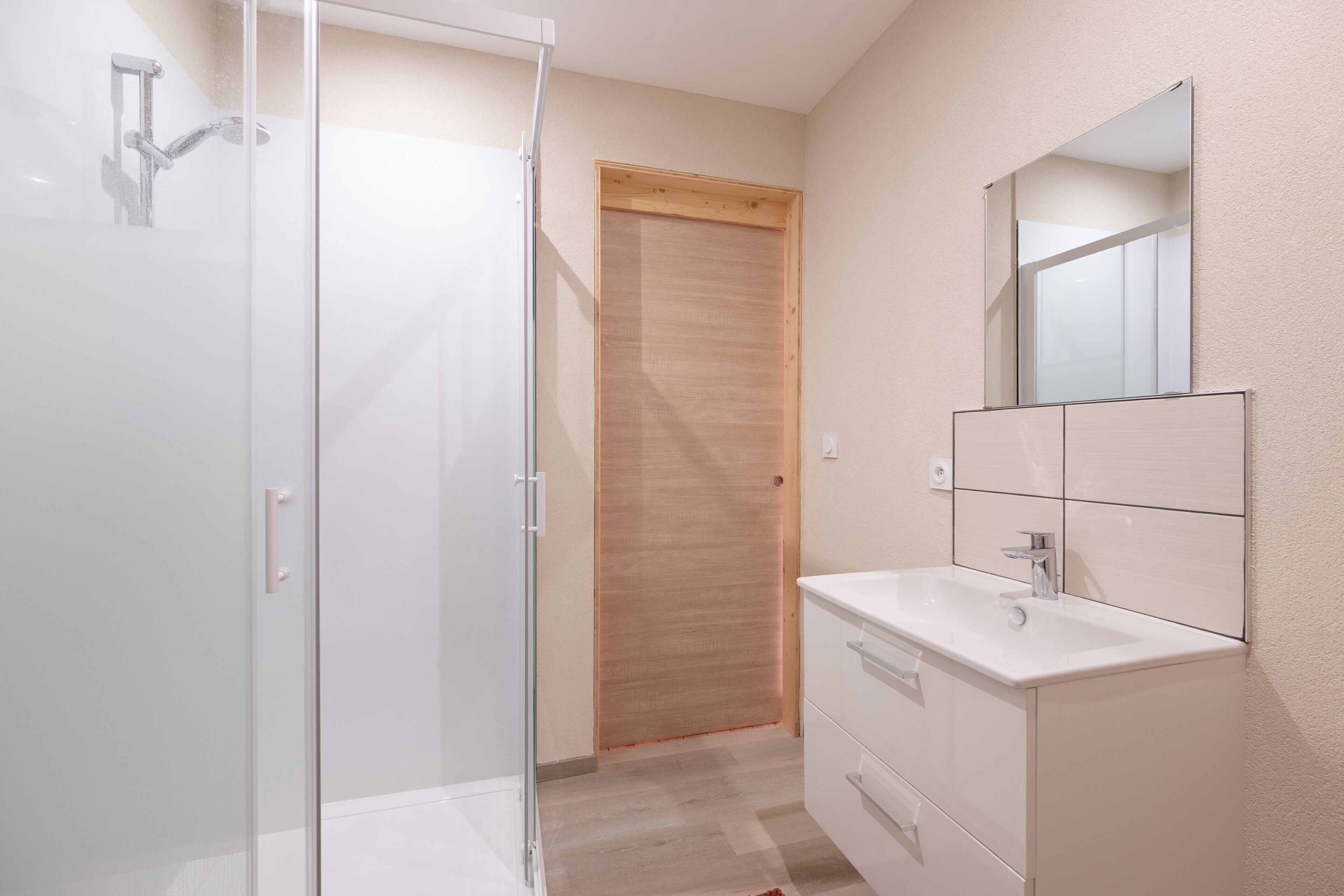 A modern bathroom with a glass shower enclosure, a wooden door, a white vanity with a mirror, and a tiled backsplash.