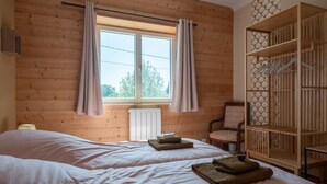 A room with wooden walls, a bed with white linens, a chair, a wooden wardrobe, and a window with curtains. - Mirabelle Room with Wi-Fi (Brû)