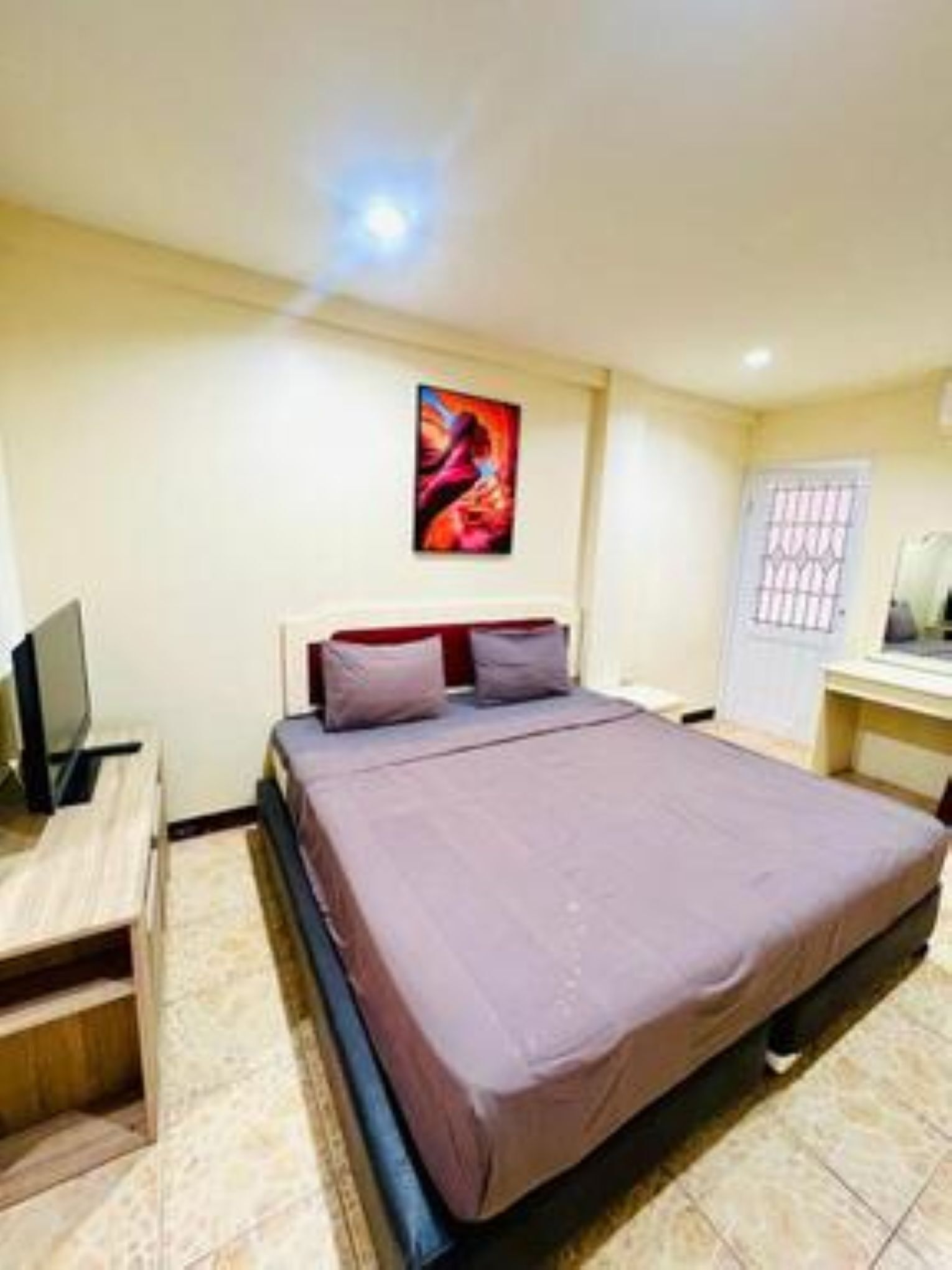 A bedroom with a bed, a desk with a computer, a door, and a painting on the wall.