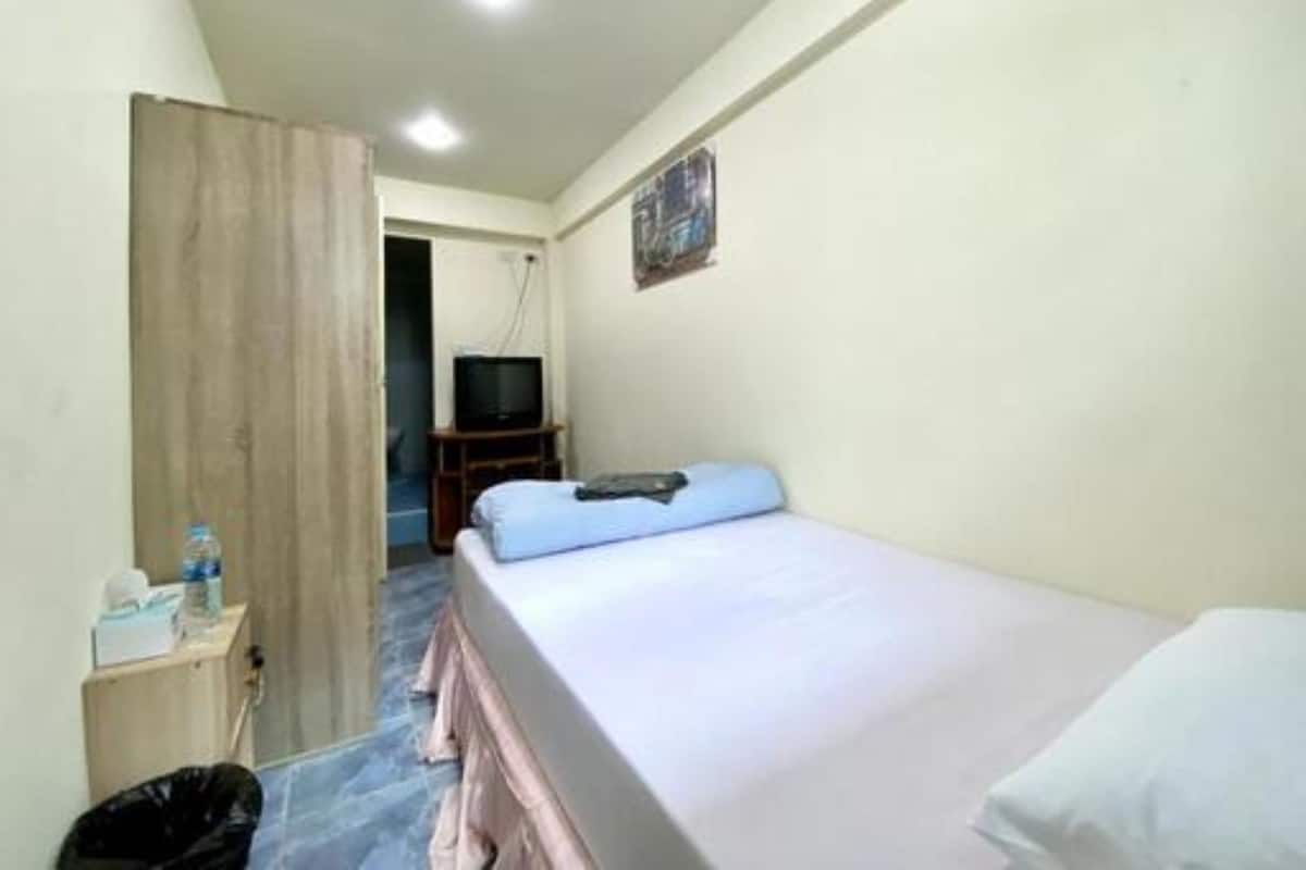 Habitación pequeña con cama individual, puerta de madera, televisor sobre un mueble de madera y una mesita con un cesto de basura.