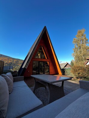 A modern, triangular wooden cabin with a large sliding glass door, a dining table, and a seating area with cushions. - Macko Cabin (Băile Tușnad)
