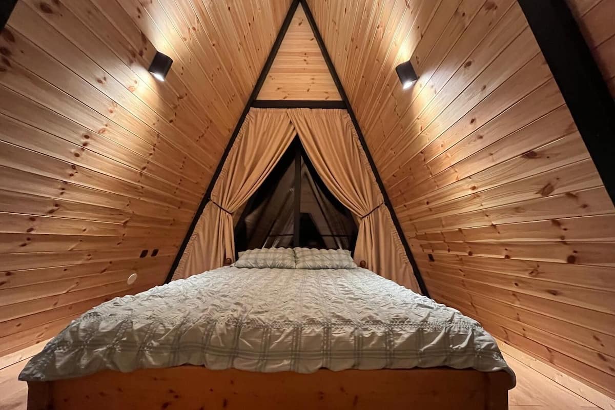 Una habitación tipo tienda de campaña de madera, con cama, paredes de madera y estructura de techo triangular.