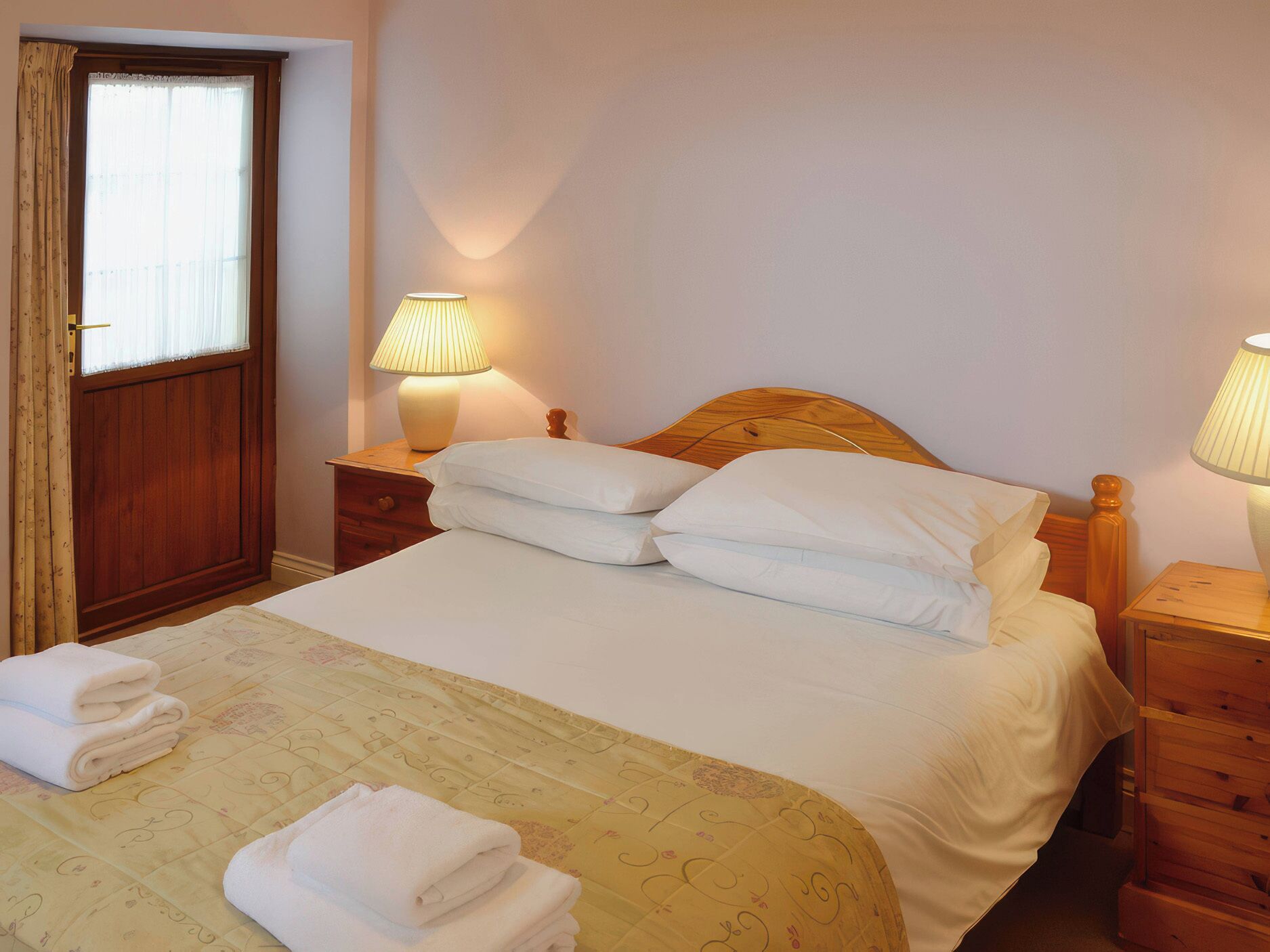 A neatly made bed with white linens, two bedside tables with lamps, a wooden headboard, and a window with curtains.