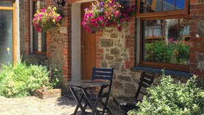 A brick building with a wooden door, hanging flower baskets, and a table with chairs outside. - Farrier Cottage (Falmouth)