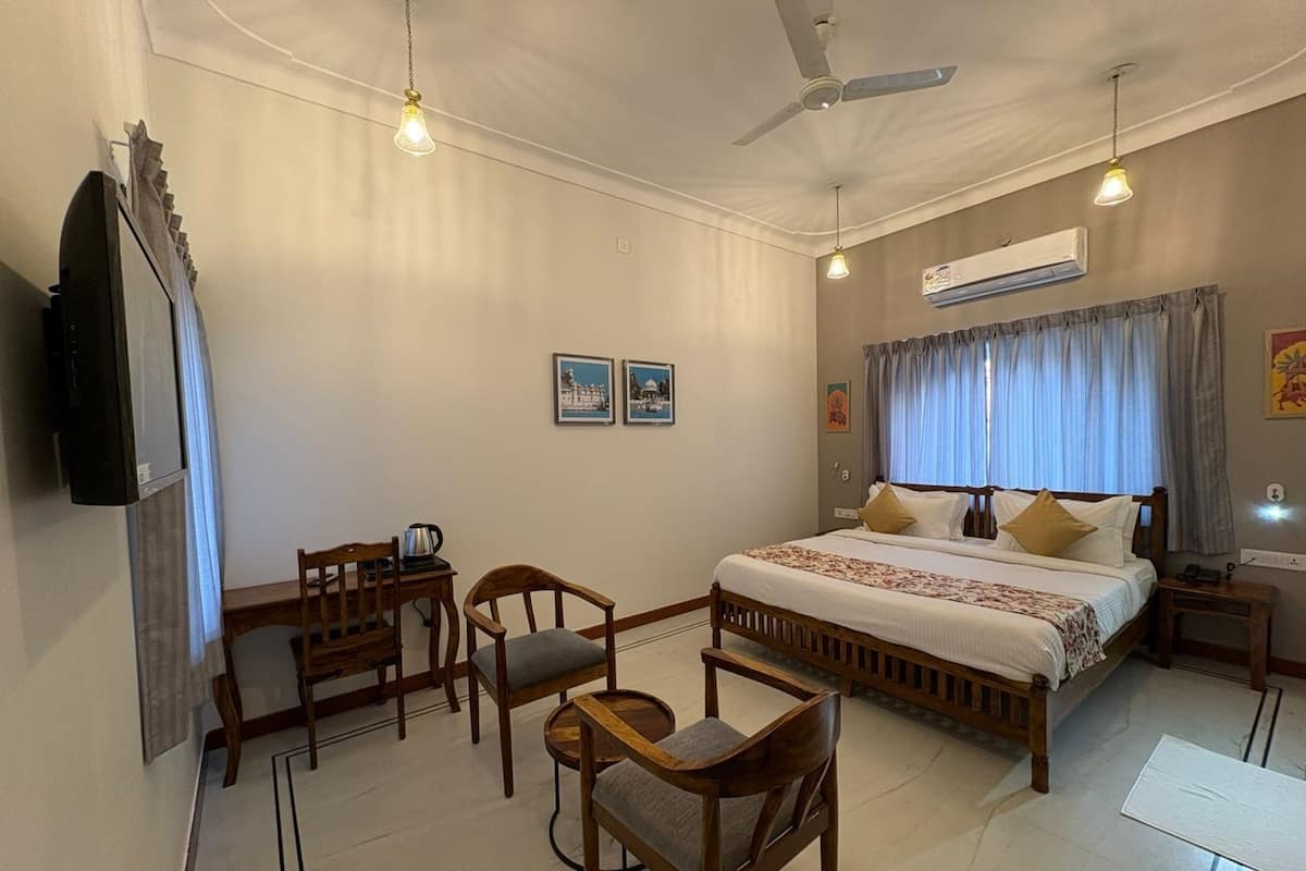 A hotel room with a bed, a desk, two chairs, a TV, and an air conditioning unit.