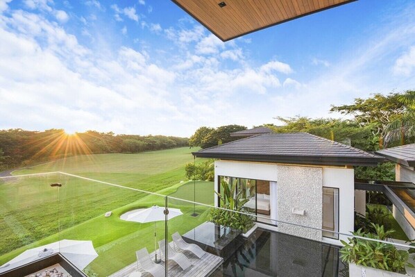 Ein modernes Haus mit einem Glasbalkon, der auf eine gepflegte Rasenfläche und einen Golfplatz blickt.