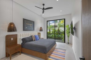 Ein modernes Schlafzimmer mit Bett, Nachttischen, Deckenventilator, großem Fenster mit Blick auf Grünflächen und einem an der Wand befestigten Fernseher.