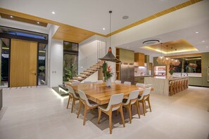 A modern dining area with a wooden table and chairs, a central hanging light fixture, and a kitchen area in the background. - Vaquero Del Mar (La Dulce Vida, Hacienda Pinilla)