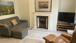 Ein Wohnzimmer mit grauem Sofa, einem braunen Ledersessel, einem Kamin, einem Fernseher und einem Gemälde von Sonnenblumen.