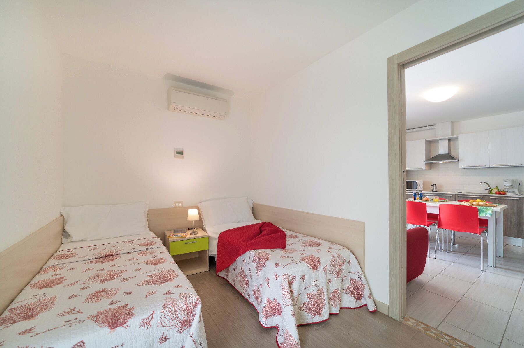A small, well-lit bedroom with two beds, a bedside table with a lamp, and a view into a kitchen area with red chairs.