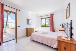 A bedroom with a bed, a wooden nightstand, a bedside table with a lamp, a window with a view, and a sliding glass door leading to an outdoor area.