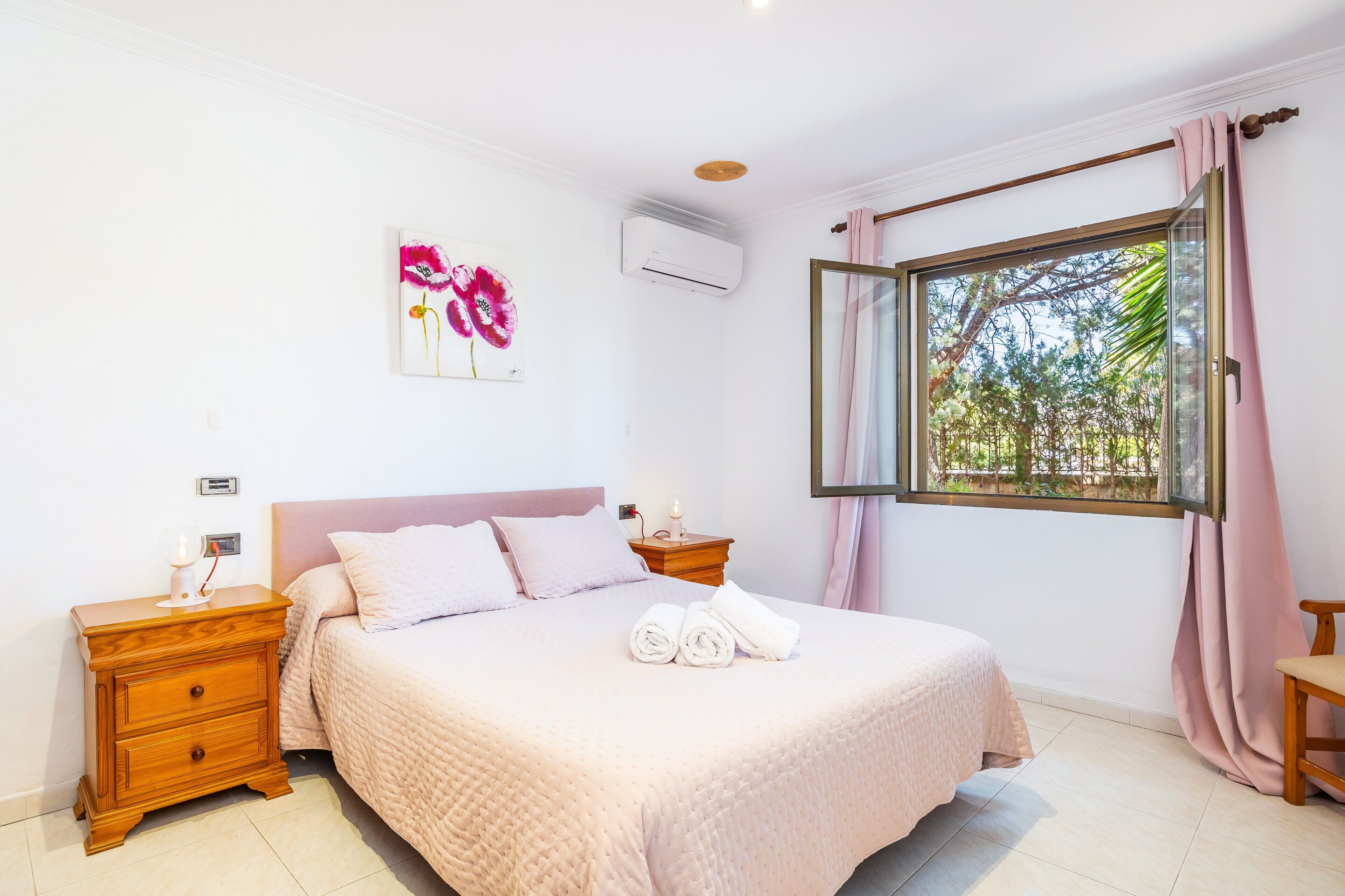A bedroom with a bed, a nightstand, a wall-mounted air conditioner, a window with curtains, and a painting.