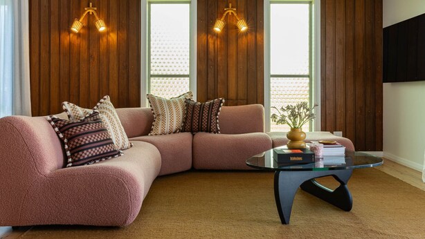 Una sala de estar con un sofá seccional rosa, dos lámparas fijadas a la pared, una mesa de centro de vidrio y un jarrón con plantas.