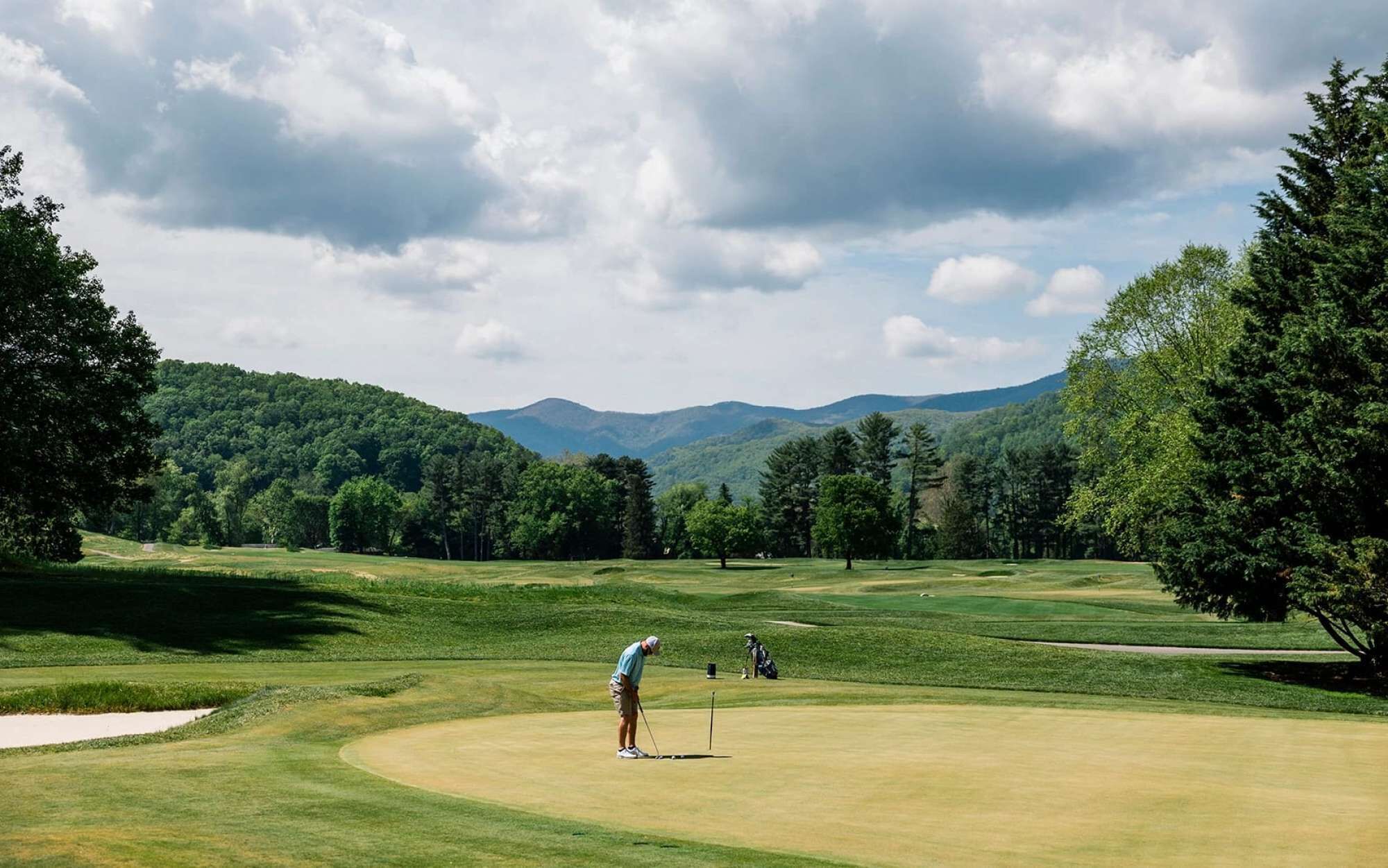 Un terrain de golf où une personne se prépare à effectuer un putt, entourée d’arbres et avec des montagnes en arrière-plan.